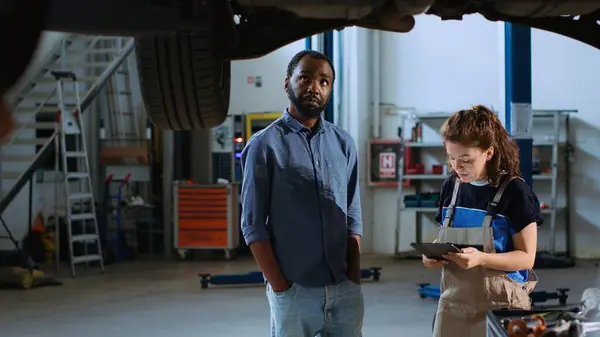 Meticulous technician in garage workplace stands with customer underneath suspended car, looking together for replacement parts. Expert helps client mend automobile using tablet to find components