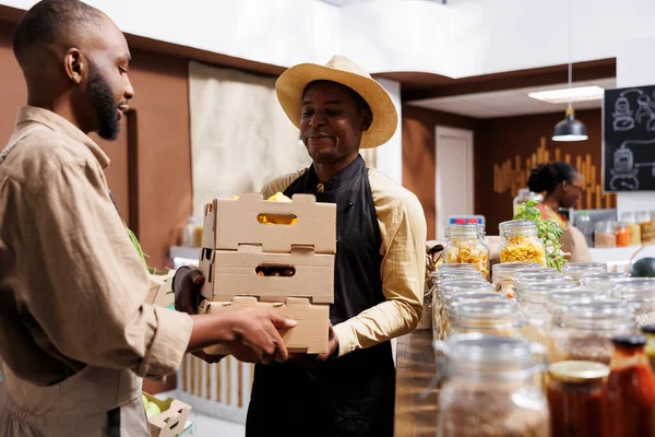 stock image Black farmer provides locally sourced bio produce to happy vendor selling it to sustainable living clients in zero waste shop. Man hands crates full of fresh natural produce to the smiling shopkeeper.