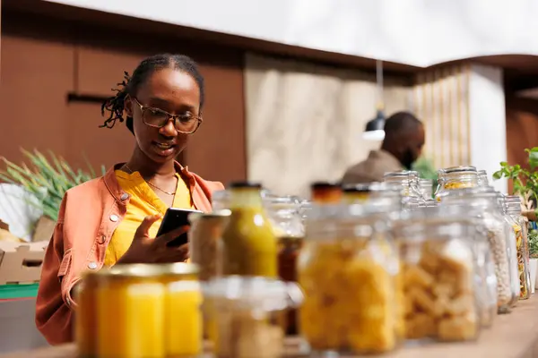 African American woman on her smartphone, surrounded by eco friendly products and reusable jars. Image shows a young black lady using her mobile device to compare products and prices.