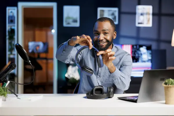 アフリカ系アメリカ人のブロガーがテクノロジー ビデオ ビデオを制作し 新たにリリースされたヘッドフォン ペアを視聴者に展示している 笑顔の男は加入者に挨拶し ワイヤレス音楽リスニングギアを提示する — ストック写真