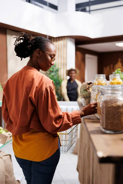 African American client shops for fresh, locally grown produce in a grocery store. They verify organic options and explore eco friendly packaging. The store offers sustainable, zero waste choices for