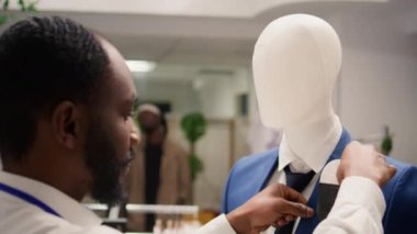 Promosyon sezonunda giyim mağazasında çalışan bir işçi manken ceketine kravat takıyor. Dükkan sahibi, moda mağazasında eski koleksiyon giysilerine cep mendili yerleştiriyor.
