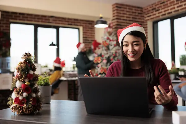Smiling employee wearing santa hat managing project in teleconference using laptop in xmas decorated office. Cheerful woman worker having online communication at workplace during new year holiday