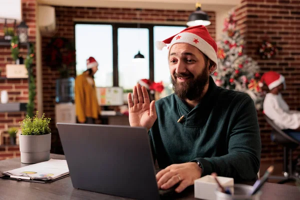 Smiling employee in santa hat waving hi while speaking at online corporate meeting using laptop. Caucasian man greeting coworkers in videocall and working in office at christmas eve