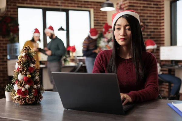 Asian woman corporate worker in santa hat working on project at winter seasonal holiday in decorated office. Company employee using typing on computer at festive coworking space