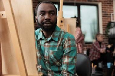 Yaratıcı hobi. Genç Afrikalı Amerikalı adam resim sehpasında resim yapıyor, yerel sanat derslerine katılıyor, çizim yoluyla yaratıcılığını ifade eden siyah adama odaklanıyor, sanat eserleri yaratıyor. Resim okuyan insanlar