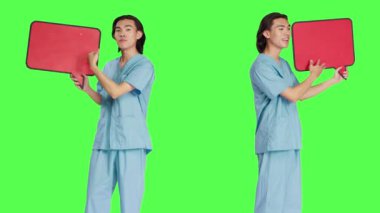 Tıp hemşiresi stüdyoda konuşma baloncuğunu yeşil perde arkaplanıyla sunuyor, karton simgeyle izole edilmiş bir kopya alanı tutuyor. Sağlık görevlisi kamerada küçük reklam panosu işareti gösteriyor..