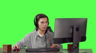 Asyalı bir oyuncu, çalışma masasında, başkalarıyla internet video oyunları oynuyor. Genç adam rol yapma oyununda eğleniyor, arkadaşlarıyla yeşil ekran şablonu üzerinden iletişim kuruyor..
