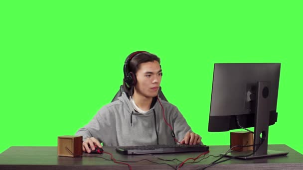 ワークステーションでPcでゲームをするアジア人 友達とオンラインビデオゲームをプレイ マルチプレイヤーコンテストで楽しんでいる若い男 グリーンスクリーン上のヘッドフォンの人々に話す — ストック動画