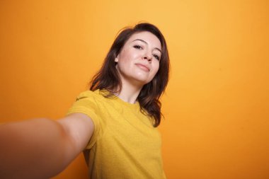 Sarı tişört giyen, izole edilmiş turuncu arka planda kamerayla selfie çeken beyaz kadın etkisinin POV 'u. Esmer kaygısız video kaydedicisi günlük bir video kaydı yaparken kameraya gülümsüyor.
