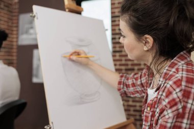 Şövalede oturan genç bir kadın basit bir vazo çiziyor. Kalem kullanıyor, öğrenci kız öğreniyor. Sanat okulunda öğrenim görürken temel şeyleri öğreniyor.