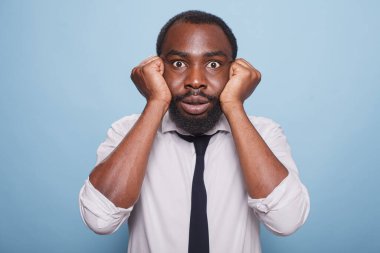 Mavi bir arkaplanda, gözleri açık sinirli bir girişimci yüzünü kapalı avuçlarında tutuyor. Afro-Amerikan bir iş adamının portresi. Yumrukları yanaklarının üzerindeydi..