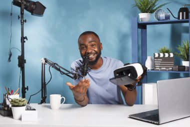 Neşeli Afro-Amerikan blogcu video sunumunda video kulaklığı tutuyor ve gösteriyor. Web içeriği yaratıcısı canlı sanal gerçeklik gözlüğü incelemesi ve mikrofon ile konuşuyor