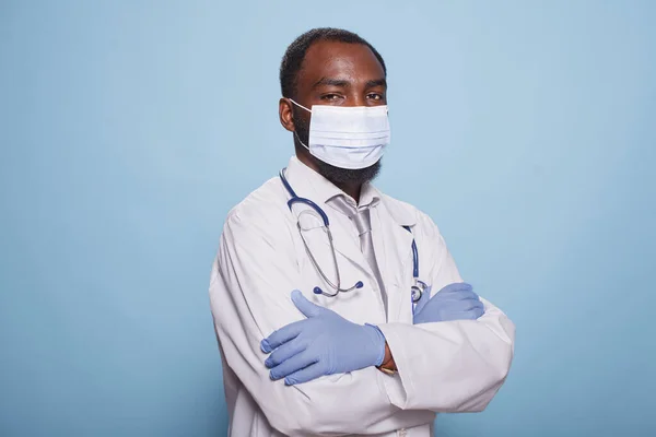 African american medic in hospital uniform wearing protective face mask and posing with his arms crossed. Portrait of black male doctor with stethoscope and medical protective gear.