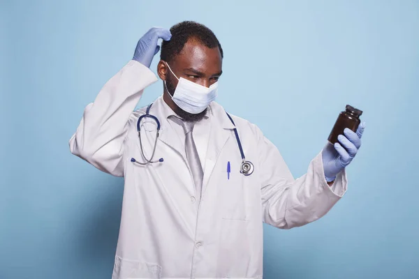 Healthcare specialist dressed in medical protective gear looking baffled at prescription pill bottle. Confused black doctor wearing face mask, stethoscope, and gloves gripping a painkiller container.