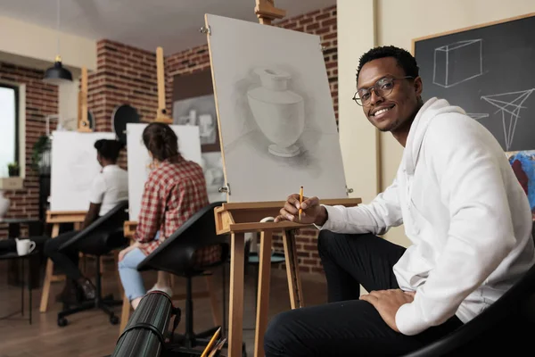 Happy African American guy sitting at easel in classroom enjoying making art during group drawing class, creative space for social connection. Smiling man learning how to draw at art workshop