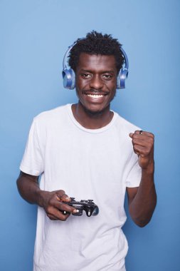 Kablosuz kulaklıklı, gülümseyen ve konsolda video oyunları oynayan heyecanlı zenci oyuncu portresi. Neşeli bir Afrikalı Amerikalı günlük giysiler içinde elinde bir kumandayla zaferi kutluyor..