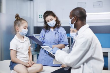 Mavi önlüklü hemşire kız hakkında bilgi içeren bir dosya dolduruyor. Yüz maskelerini hasta, annesi ve çok kültürlü sağlık personeli takıyordu. Profesyonellik atmosferi.
