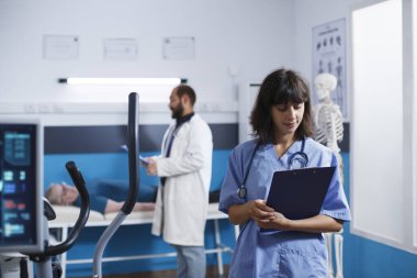 Pratisyen hemşirenin durduğu portresi olan fizyoterapi ofisi evraklarını kontrol ediyor. Tıp hekimi hastalara fiziksel rehabilitasyon egzersizleri için yardım etmeye hazırlanıyor..