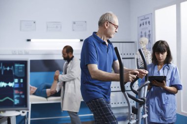 Yaşlı erkek hasta, mavi önlüklü hemşirenin desteğiyle sabit bisiklet üzerinde fizyoterapi ile meşgul. Dijital tabletli tıp asistanı son sınıf öğrencisine fizik tedavisinde yardım ediyor..