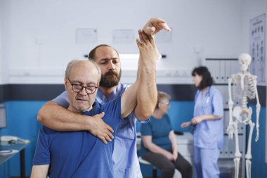 Tıp hemşiresi yaşlı birinin kolunu yükselterek kaslarını esnetiyor ve fiziksel yaraları tedavi ediyor. Fizyoterapi konsültasyonunda, erkek osteopat kıdemli hastalarla hareket etme egzersizleri yapar..