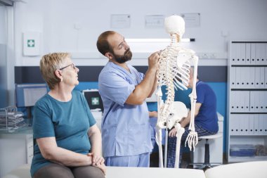 Hemşire ve emekli hasta osteopatik muayene için insan iskeletinde omuriliğe bakıyor. Sağlık hizmetlerinden sorumlu yaşlı kadına teşhisi açıklamak için omurga kemiklerine işaret eden tıbbi asistan..
