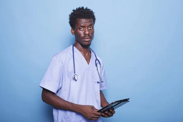Male healthcare professional with stethoscope stands in profile, holding a digital tablet. African American nurse wears scrubs and carries modern technology to address COVID-19 concerns.