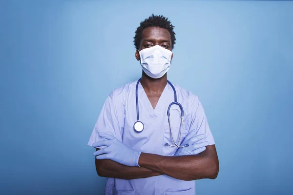 Portrait of a black nurse practitioner posing with his arms crossed on an isolated background. Looking at the camera is an African American man wearing gloves, a face mask, and blue scrubs.