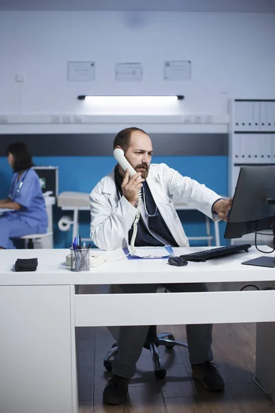 In medical office, male physician is using a computer for medical research and chatting on a wired phone. Caucasian man in lab coat using landline phone to speak with hospital receptionist.