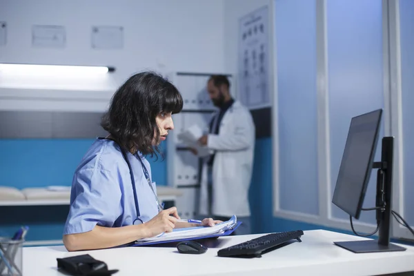 Professional medic looking at computer screen and medical files for healthcare. Nurse reviewing information on pc monitor and documents for assistance and support, doing overtime work.