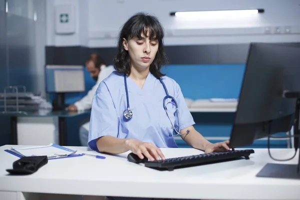Young medical assistant works on computer at desk. Working late in the office, nurse in uniform examines a monitor for paperwork and a healthcare checkup. Healthcare with modern technology.