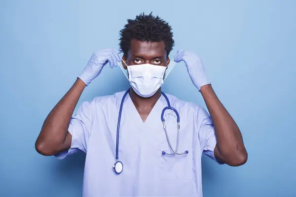 Portrait of healthcare specialist putting mask on face for protection against covid 19 pandemic. Man working as nurse wearing uniform, stethoscope and gloves while looking at camera.