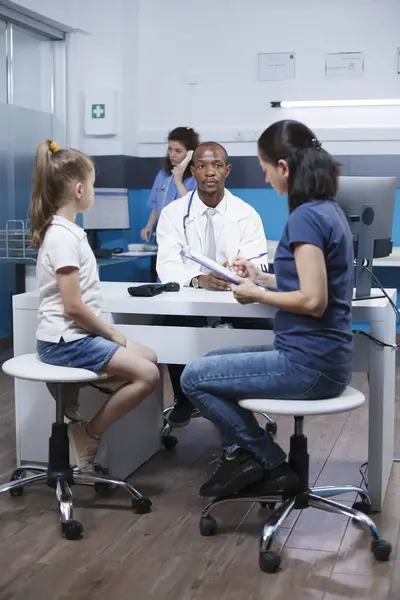 病院のオフィスにいる白人女性と娘が医学検査のために書類を記入した アフリカ系アメリカ人医師は 女性がクリニックで書類を完成するのを待つ — ストック写真