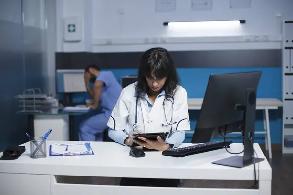 Front-view of female physician using a tablet in clinic office to analyze healthcare data. Caucasian doctor in lab coat, concentrated on reviewing patient medical information on her digital device.