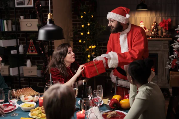 Santa claus giving presents to people at festive dinner, spreading christmas spirit and enjoying december holiday. Man dressed in costume and hat offering gifts to friends and family.