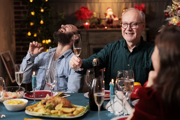 Old adult giving toast with raised glass of champagne at christmas eve celebration to enjoy winter holiday with friends and family. Grandpa making speech around the table, sparkling wine.