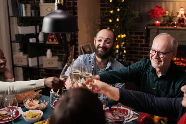 Diverse people clinking wine glasses at christmas eve dinner, celebrating holiday event with alcohol and homemade food. Friends and family saying cheers with drinks at table, cozy decorations.