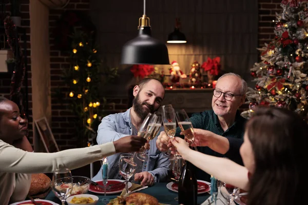 Diverse persons clinking alcohol glasses at christmas eve dinner, celebrating holiday tradition with wine and homemade meal. Friends and family saying cheers with drinks at table, cozy ornaments.