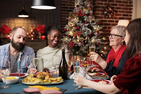 Grandma makes toast at dinner table with friends and family, enjoying christmas eve festivity at home. Senior woman giving speech with raised wine glasses for cheers, winter celebration.