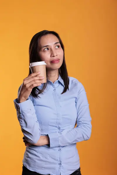 满意的模型在黄色背景下拿着一杯咖啡 在镜头前喝着热饮料 在演播室摆姿势时 微笑的亚洲女人一边喝着点心 一边喝着浓咖啡 — 图库照片
