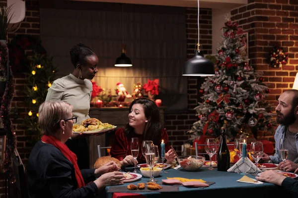 Cheerful people at festive dinner, meeting to celebrate christmas eve together with family. Diverse men and women serving meal and drinking wine glasses, feeling joyful during winter season.