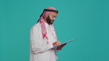 Arap biri mavi arkaplanda duran dosyalara mektup yazıyor, yeni bir program üzerinde çalışıyor ve notlar alıyor. İslami giyinmiş bir adam stüdyoda evrak işleri için kontrol listesine bakıyor..