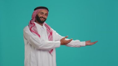 Geleneksel cübbe ve ghutra giymiş Müslüman yetişkinler, taraflarını elleriyle sunuyor ve stüdyo çekimi için poz veriyorlar. Arap neşeli adam pazarlama reklamını bir kenara bırakarak bir şeyleri destekliyor..