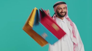 Stüdyoda alışveriş torbaları tutan mutlu Arap, indirimli ürünler aldıktan sonra keyifli hissediyor. Ortadoğulu, perakende mağazasında indirimli ürün satın alıyor. Renkli kağıt poşetler..