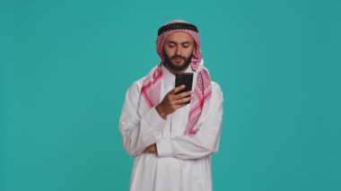 Geleneksel kıyafet giymiş Müslüman bir adam akıllı telefon kullanıyor, çevrimiçi sosyal medya konuşmalarında kamera önünde yer alıyor. Orta Doğulu adam, başörtüsü ve cübbe takıyor, internette geziniyor..