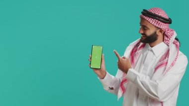 Orta Doğulu adam akıllı telefondaki yeşil ekran ekranını işaret ederek mobil cihaz ekranında izole edilmiş telif alanı şablonu gösteriyor. İslami kostümlü genç adam boş kromakey gösteriyor..
