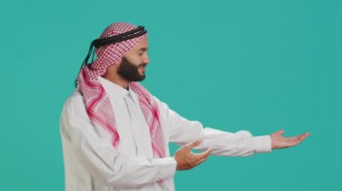 Geleneksel bornoz ve ghutra giyen Müslüman adam elleriyle hareket ediyor ve stüdyo çekimi için poz veriyor. Ürünü tanıtan ve pazarlama reklamını bir kenara bırakan Arap gülümseyen kişi.