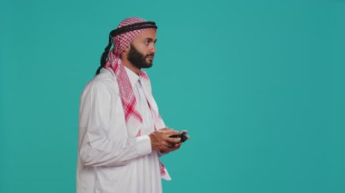 Modern adam kumandayla oyun oynuyor, mavi stüdyo arka planında video oyunu yarışmasıyla eğleniyor. Geleneksel Arap kıyafeti giyen İslamcı oyuncu turnuvada yarışıyor.