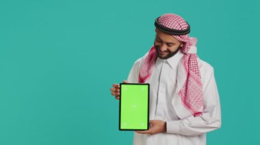 Arap adam stüdyo arka planında yeşil ekran sunuyor, boş bir telif alanı maketi ekranı sergiliyor. İslami kostümlü genç, izole edilmiş kromatonlu tablet tutuyor..
