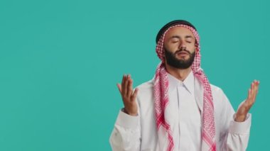 Kamerada, Arap kişi mavi arka plandaki kutsal uygulamalara güvenerek Allah 'ı arıyor. Orta Doğulu adam meditasyonla Tanrı 'yı onurlandırıyor, İslam dinine bağlılığını gösteriyor..
